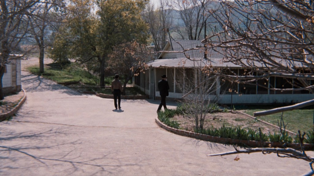 Two figures walking along a dirt path towards a house surrounded by trees and greenery.