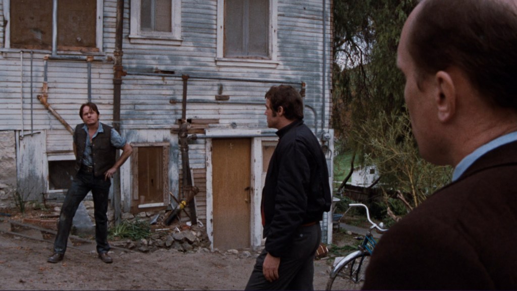A man stands outside an old, weathered house, looking at two other men who are facing him. The scene includes a bicycle near the house.