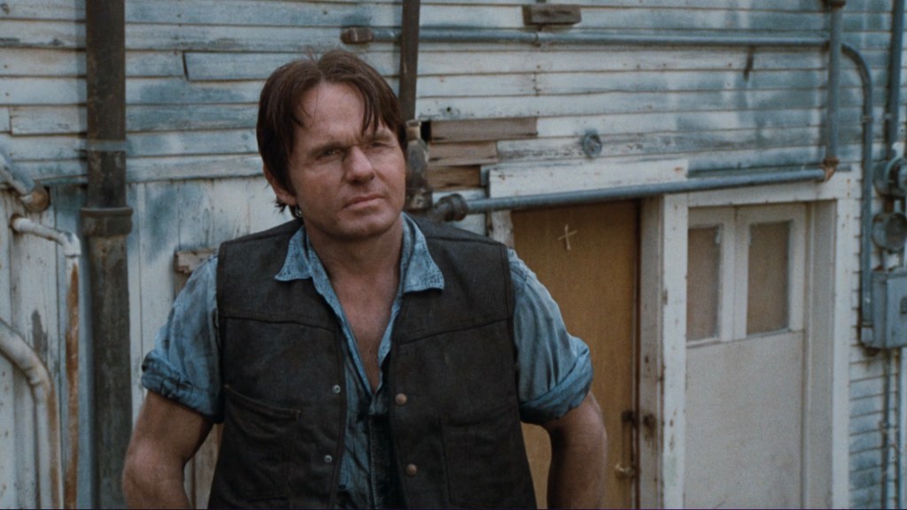 A man with medium-length hair wearing a black vest and a blue shirt stands in front of a weathered building with peeling paint and a door.