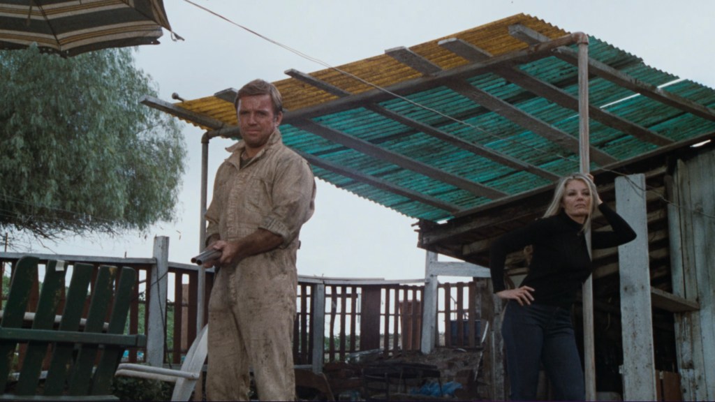 A man in a dirty jumpsuit holding a gun stands on a porch while a woman in a black outfit leans against a post, with a colorful canopy overhead.