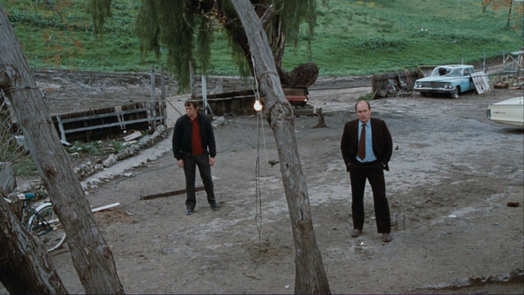 Two men standing in a barren, outdoor area with sparse vegetation and abandoned vehicles. One man wears a black jacket and the other a suit.