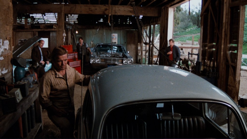 A mechanic working on a silver car inside a rustic garage, surrounded by three other men observing. Various tools and car parts are visible in the background, alongside another classic car.