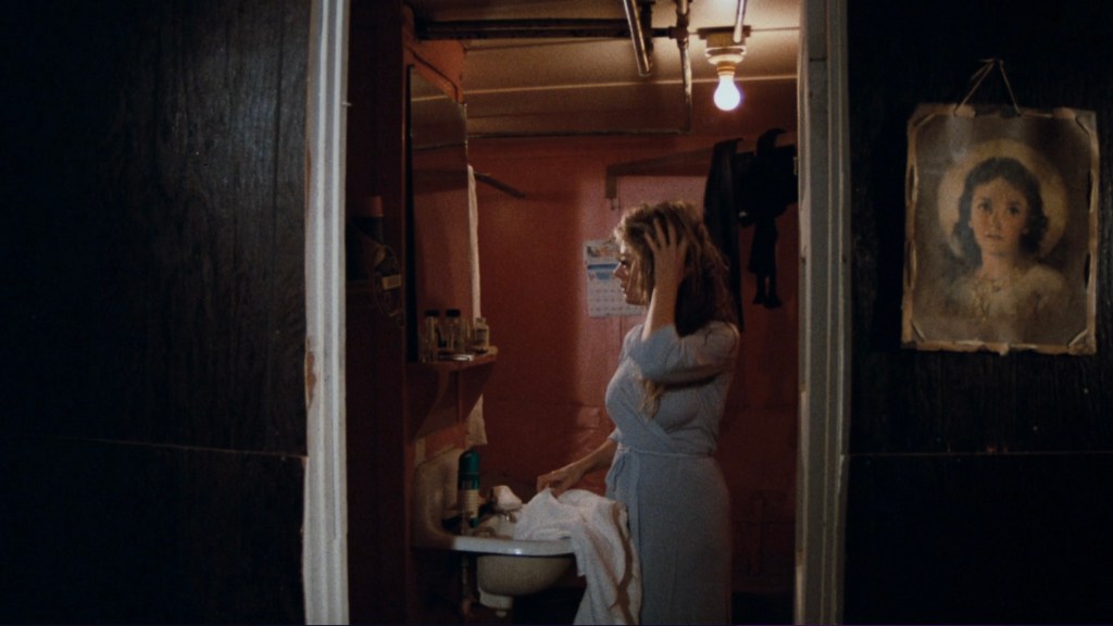 A woman with long hair is standing in a dimly lit bathroom, looking at herself in the mirror while adjusting her hair. The walls are painted pink, and there is a vintage portrait of a girl hanging on the wall.