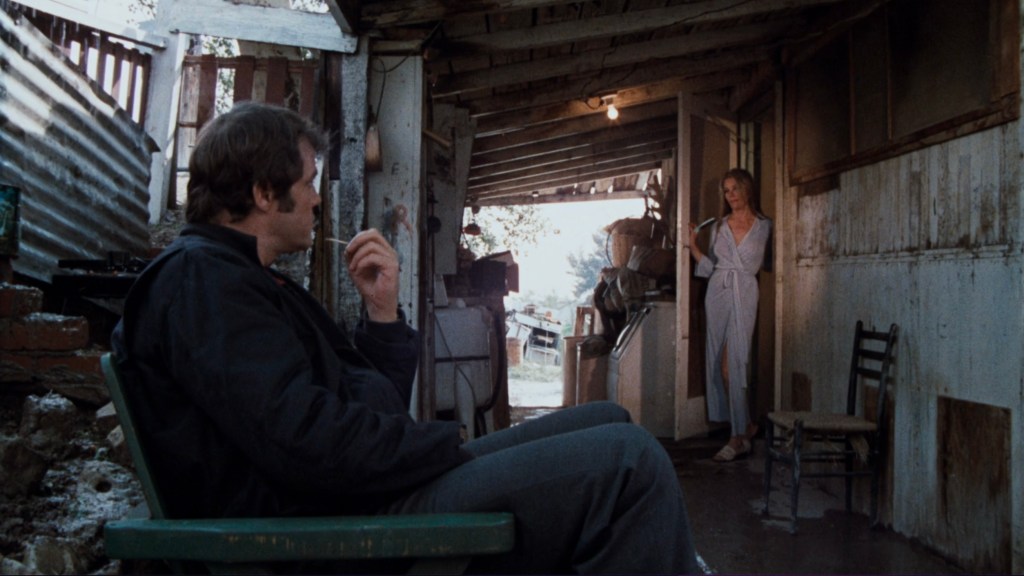 A man sitting in a chair outside a rustic building, looking at a woman standing in a doorway wearing a light-colored robe.