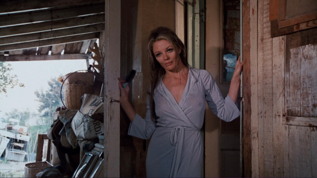 A woman in a light robe stands in a doorway, looking directly at the camera amidst a cluttered interior.