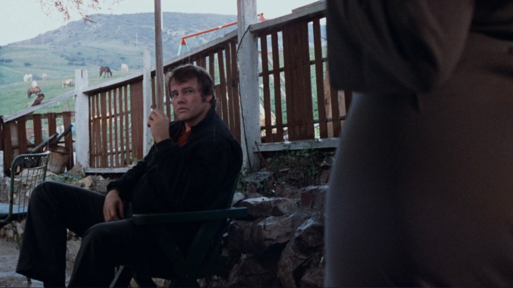 A man sitting on a chair in a rural setting, holding a cigarette and looking towards a woman partially visible in the foreground, with a backdrop of horses and grass hills.