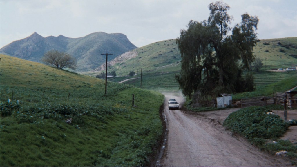A dirt road winding through lush green hills with mountains in the background and a car driving along the path.