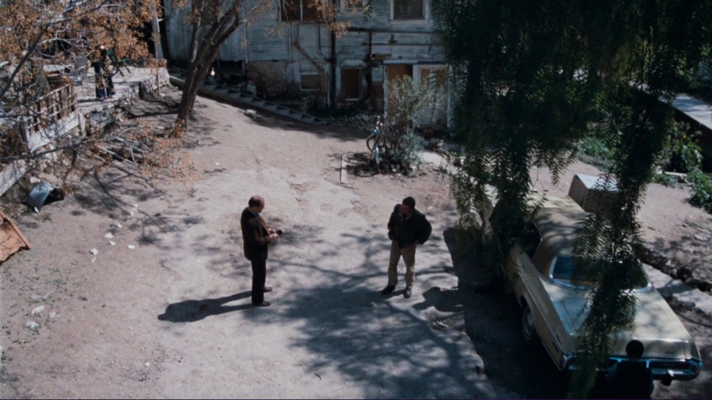Two men conversing in a dirt yard with an old car parked nearby, surrounded by trees and a vacant building.