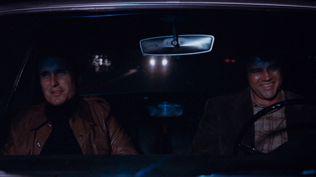 Two men sitting in a car at night, one smiling and the other looking serious, with headlights visible in the background.