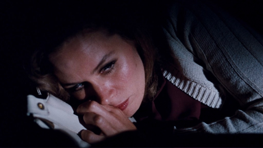 A close-up of a woman lying down, her face showing concern as she rests her head on a white object in a dark setting.