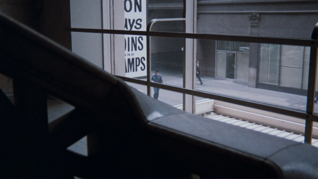 View from inside a building looking out through a window onto a city street, with two people walking by and a sign visible that reads 'ON COINS AND STAMPS.'