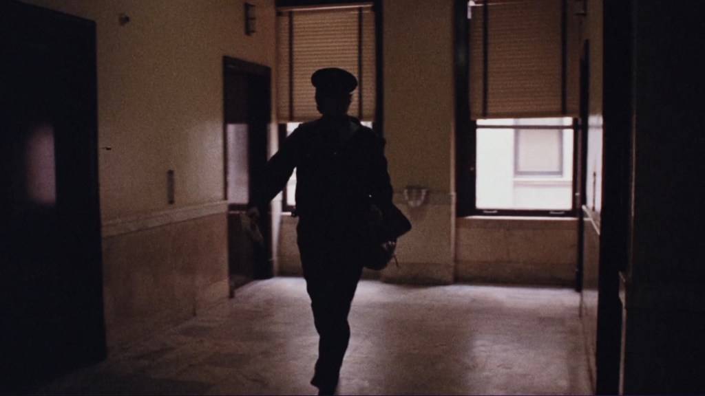 A silhouette of a person in a dark hallway, walking toward a doorway, with window blinds partially closed.