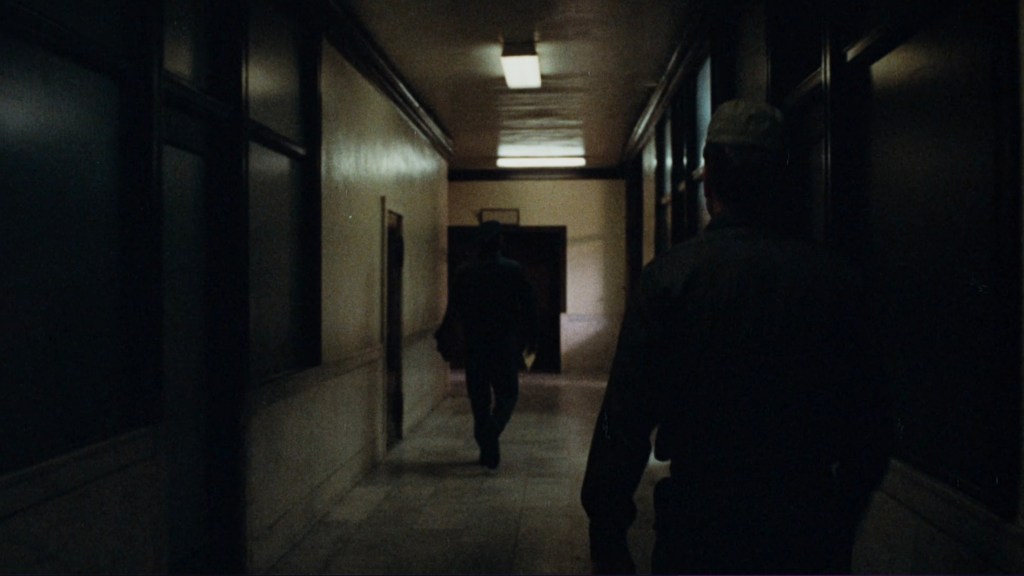 A dimly lit hallway with two figures walking, one approaching from the background and another slightly ahead, with doors on either side.