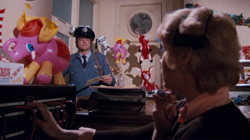 A police officer stands in an office filled with colorful inflatable toys, including a pink elephant and various other whimsical characters. A woman at a desk is holding a microphone, engaged in conversation while operating a switchboard.