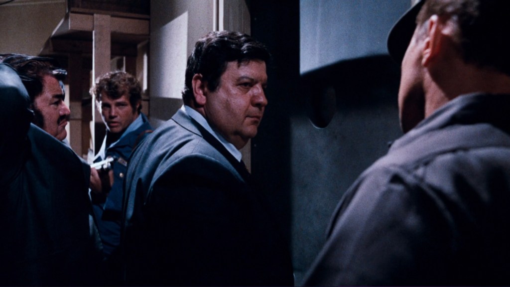 A tense scene featuring several men in formal and casual attire, engaged in serious conversation in a dimly lit corridor.