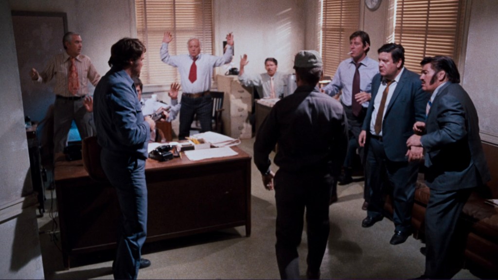 A tense scene depicting several men in a room, some with their hands raised, appearing alarmed or surprised. A man in a uniform stands in front of them while others are positioned around the room, including a desk with papers and a phone.