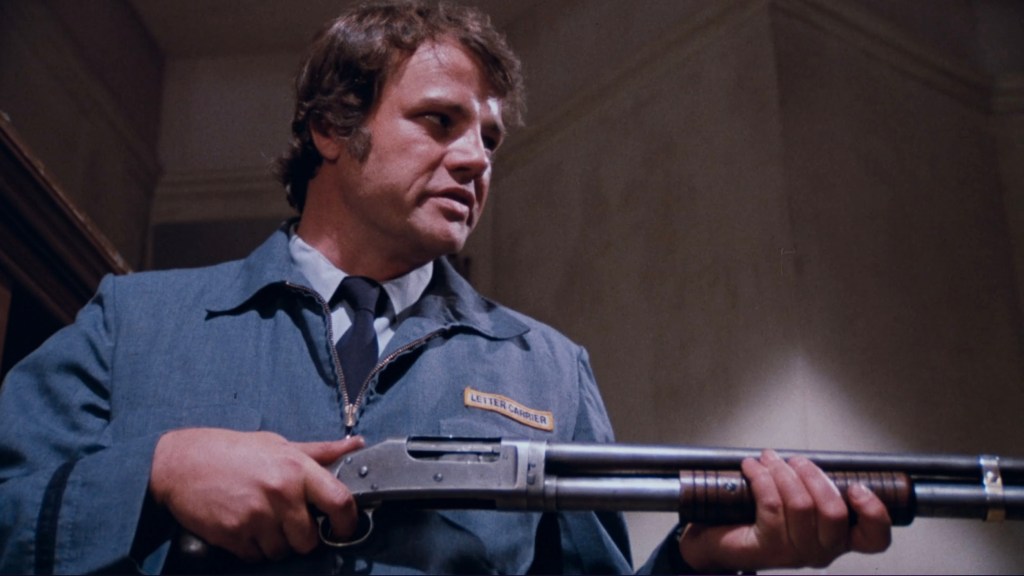 A man in a blue uniform holds a shotgun, standing in a dimly lit room.
