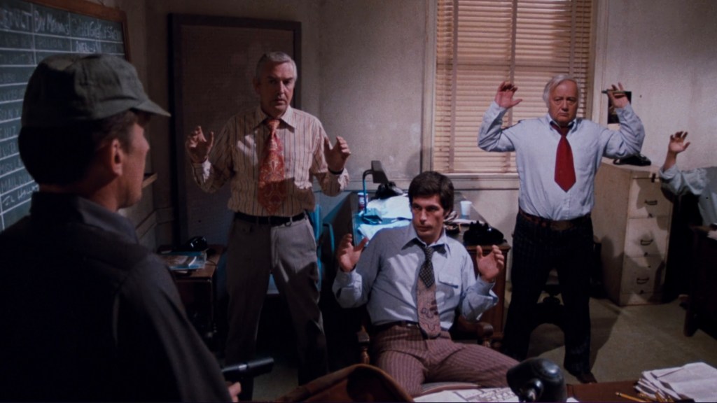 A tense scene in an office where three men are seated with their hands raised, while a fourth man, dressed in a military-style outfit, confronts them with a gun.