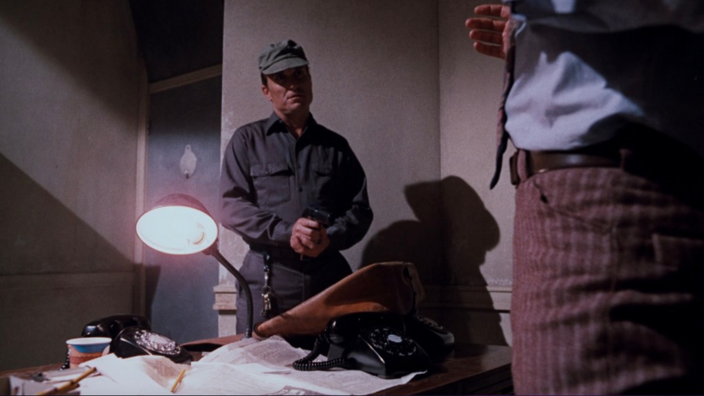 A tense scene in a dimly lit room featuring a man in military attire holding a firearm, while another man in formal clothing stands nearby. A table with a lamp, documents, and rotary phones is visible.