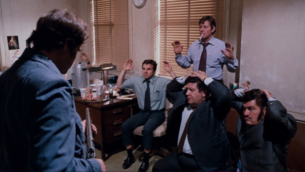 A tense scene in an office with a group of three men sitting against the wall with their hands raised, while another man stands facing them, holding a weapon. The room is furnished with a desk, a few chairs, and a window with blinds.