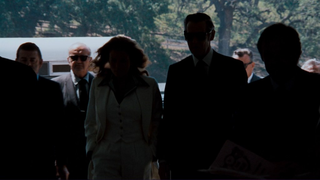 Silhouetted figures of men and a woman in formal attire entering a building, with bright light in the background.