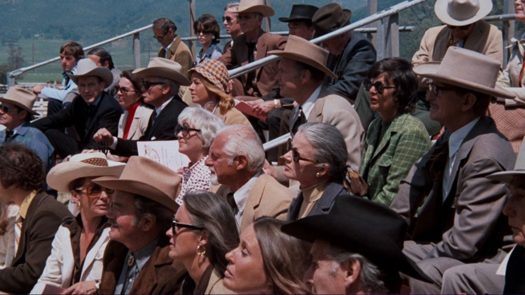 A diverse group of people seated in an outdoor setting, many wearing hats and dressed in formal attire, attending an event.