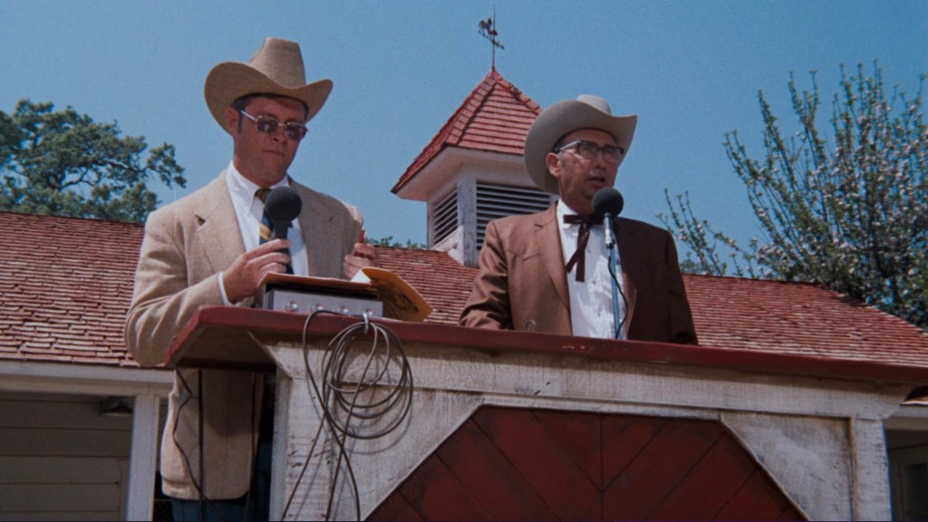 Two men in cowboy hats stand at a podium, speaking into microphones, against a backdrop of a red-roofed building and trees.