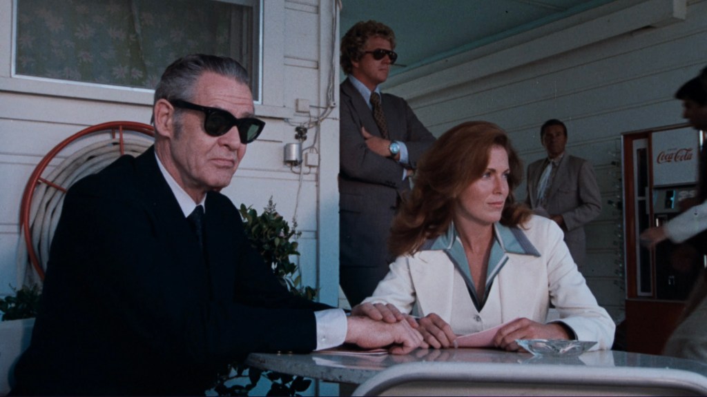 A group of people seated at an outdoor table. A man in a dark suit and sunglasses sits beside a woman in a white blazer, both looking serious. In the background, two other men stand, one wearing sunglasses and a suit, and a vending machine is visible.