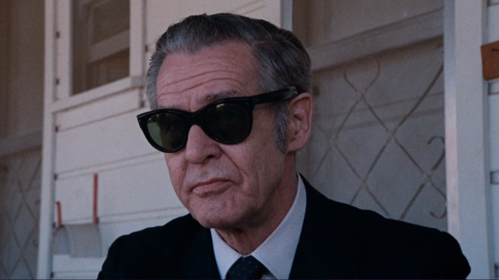 Portrait of an older man wearing dark sunglasses and a suit, sitting outdoors with a neutral expression.