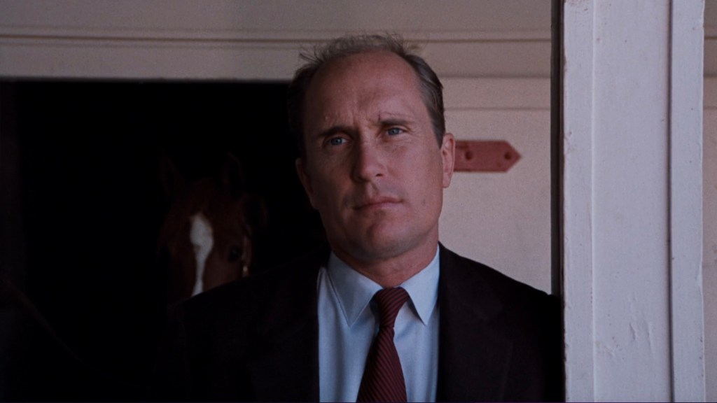 Close-up of a man in a suit with a serious expression, standing by a doorway. A horse's head is partially visible in the background.