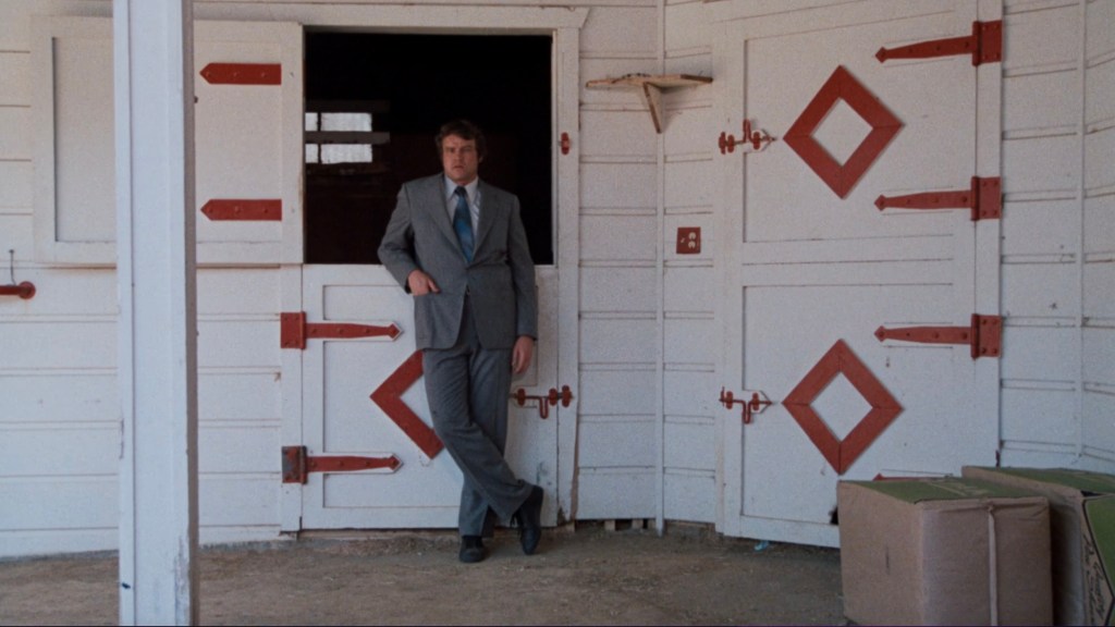 A man in a gray suit leans against a barn door, with red accents on the door and barn structure.