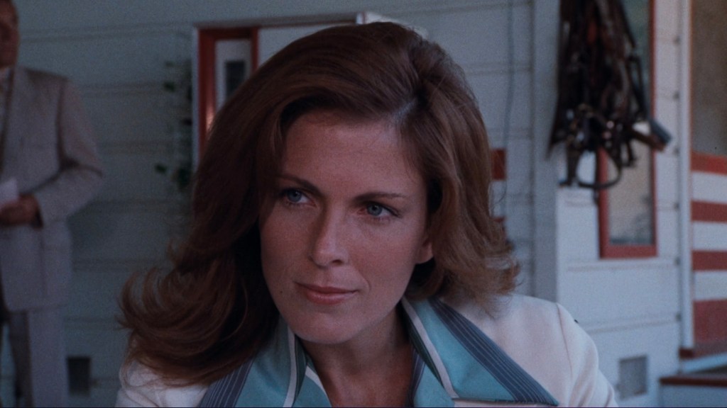 Close-up of a woman with wavy hair, wearing a light-colored jacket with a blue collar, looking intently towards the camera, with a blurred figure in the background.