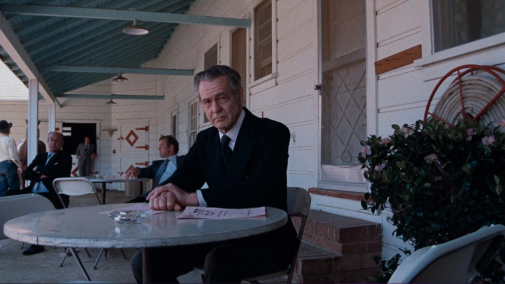 A man in a black suit sitting at a round table on a porch, with other people seated nearby and a house in the background.