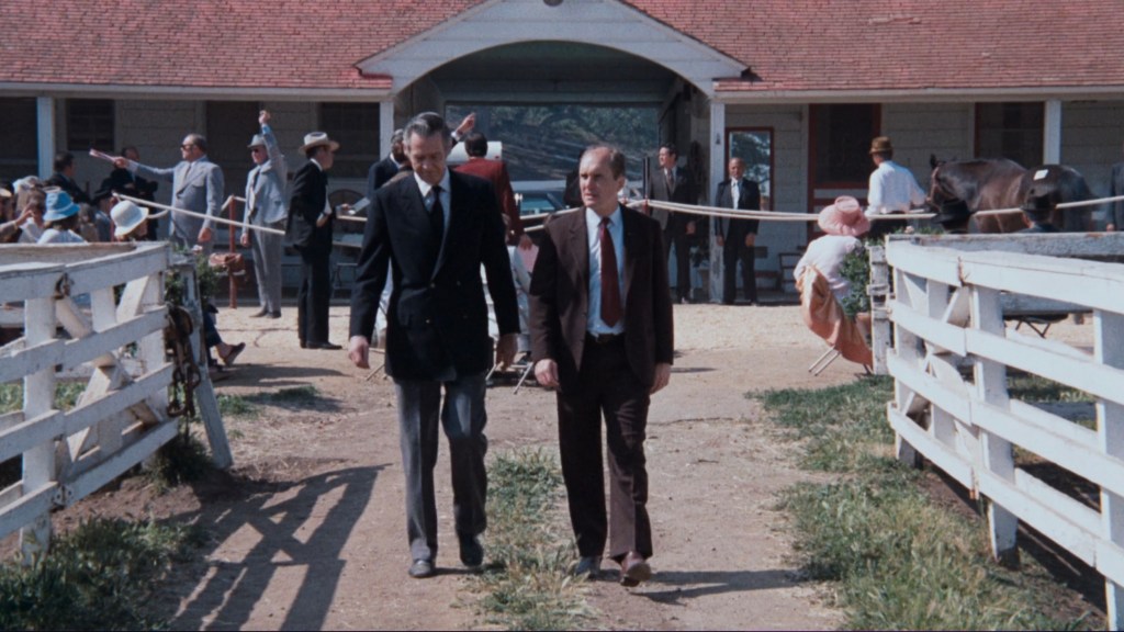 Two men in business attire walking along a path at a horse racing facility, with a crowd of people in the background and horses visible nearby.
