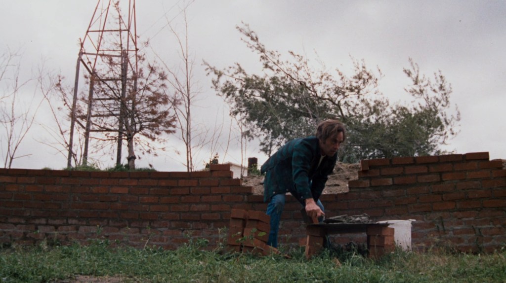 A man in a green jacket working on a brick wall construction outdoors, with a metal tower and trees in the background.