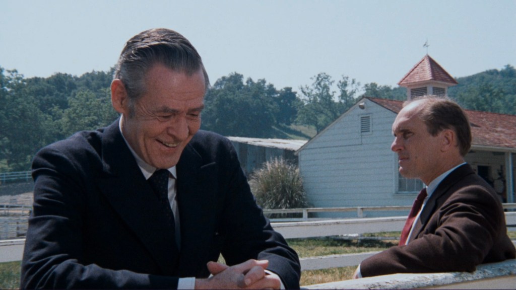 Two men in formal attire engaged in conversation outdoors, with a rural background featuring a white fence and a building.