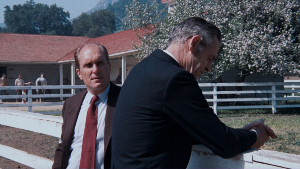 Two men in formal attire stand by a white fence at a ranch, with horses and a building in the background.