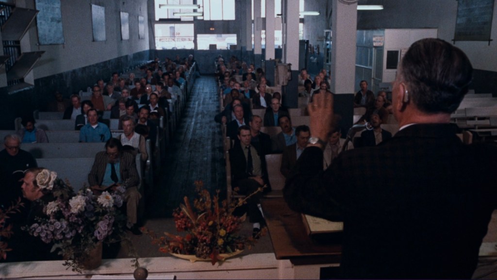 A speaker stands at a podium addressing a large audience seated in a spacious, sparsely decorated room.