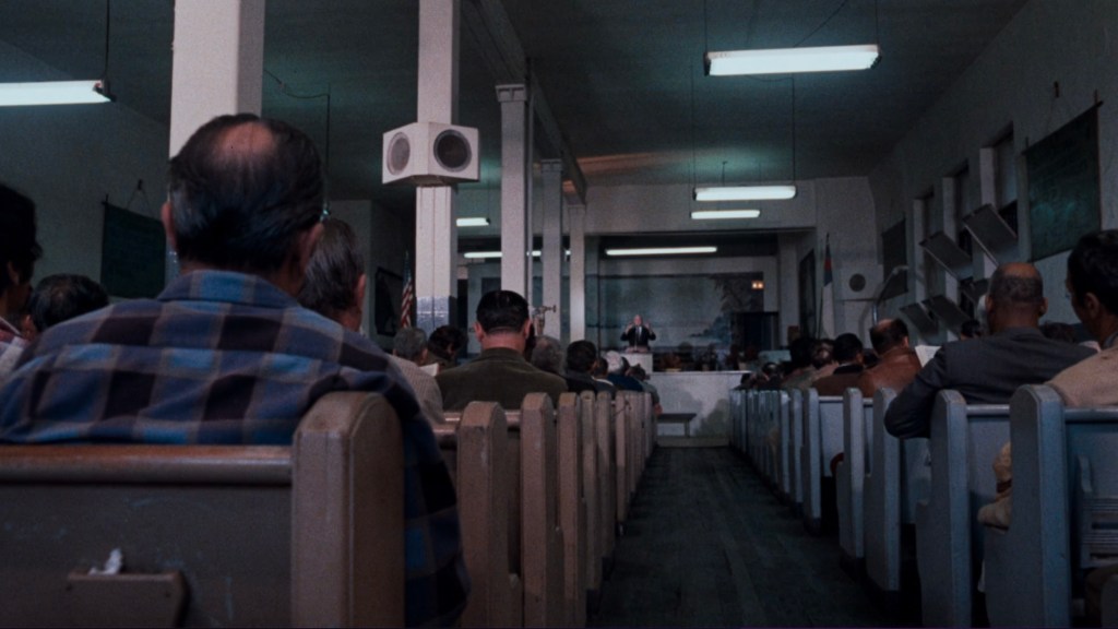 Interior view of a church-like setting with rows of wooden pews filled with people, facing a speaker at the front.