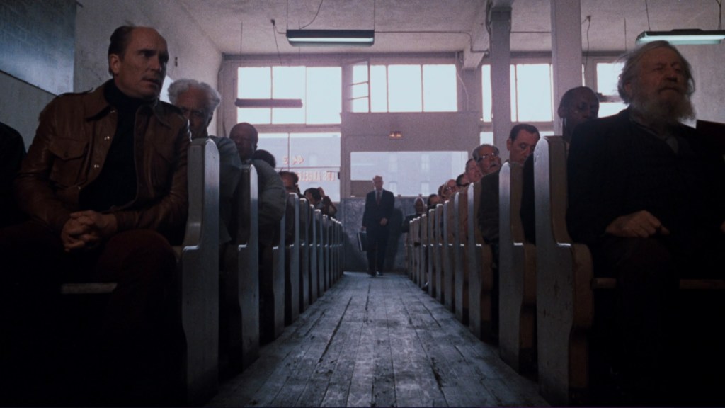 A perspective view of an empty church-like interior with wooden pews, showing a row of seated, elderly men facing the camera. In the distance, a man in a suit walks towards the viewer, illuminated by natural light coming from large windows.