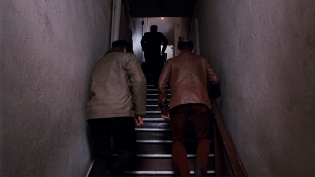 Two men ascending a staircase, with a third figure visible at the top in a dark silhouette.