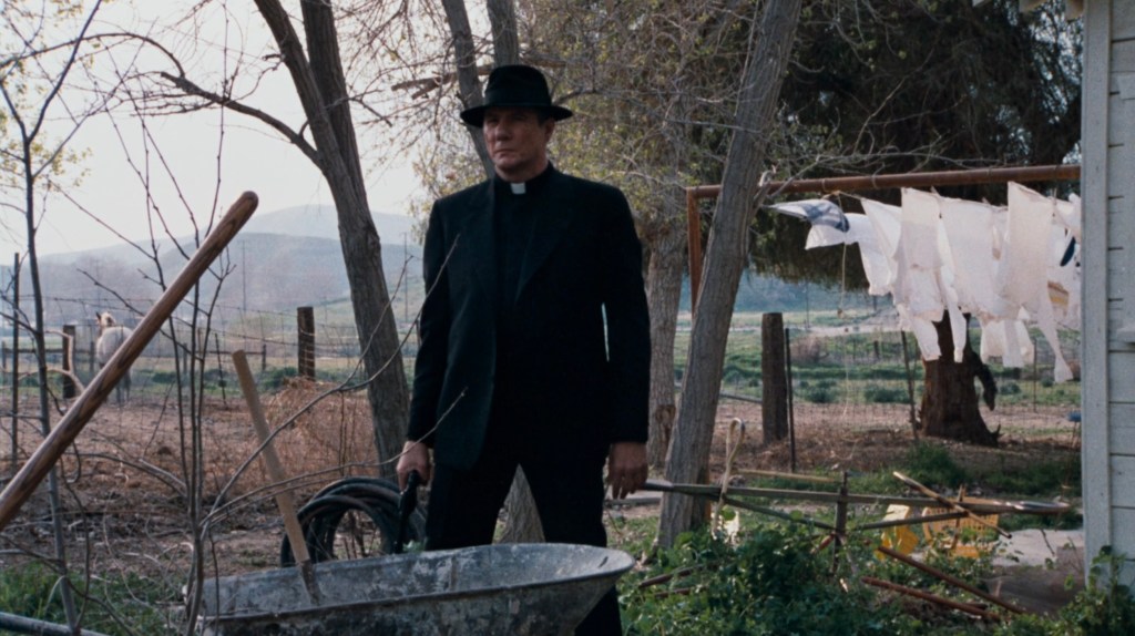 A man in a black outfit and hat stands in a rural outdoor setting, holding a tool while surrounded by trees and clothes hanging on a line.