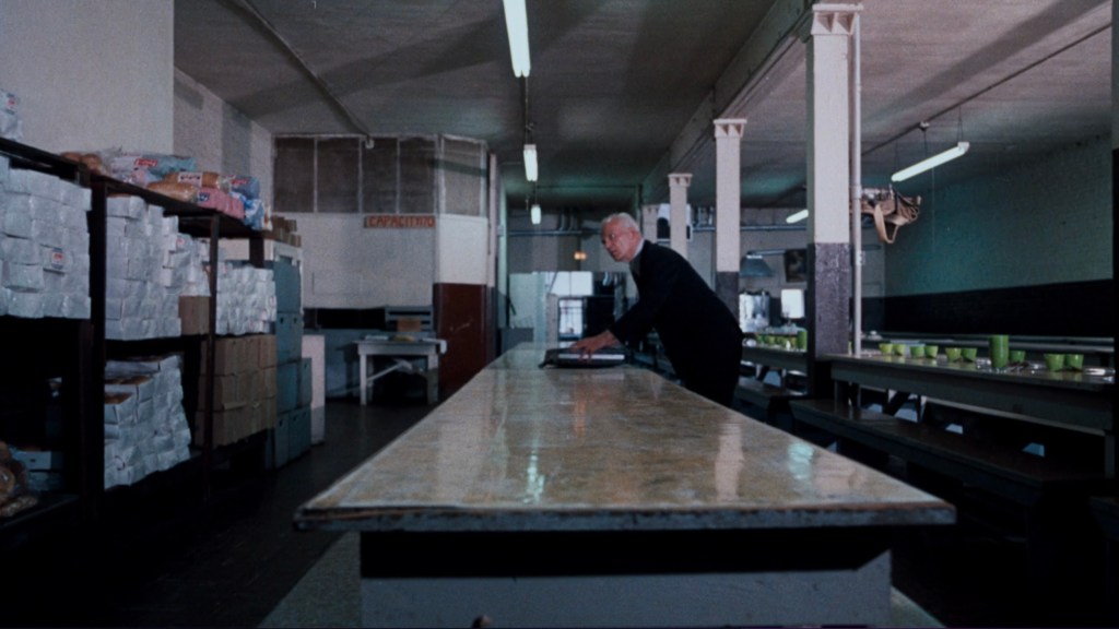 An empty dining hall with long tables and shelves filled with boxes, featuring a man in a black outfit leaning over a table.