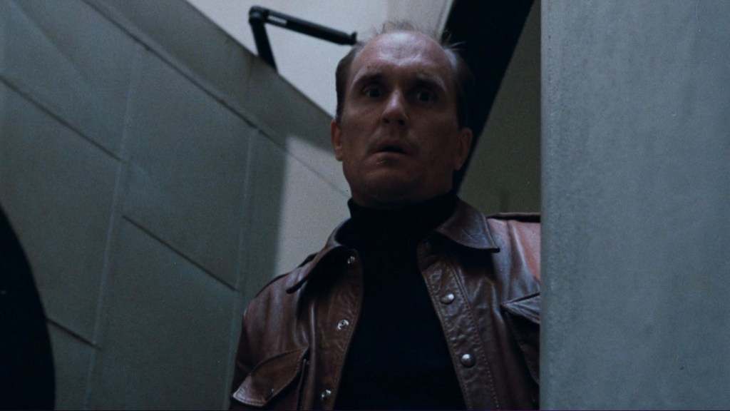 A man standing in a hallway, wearing a brown leather jacket and a black turtleneck, with a surprised or worried expression on his face.