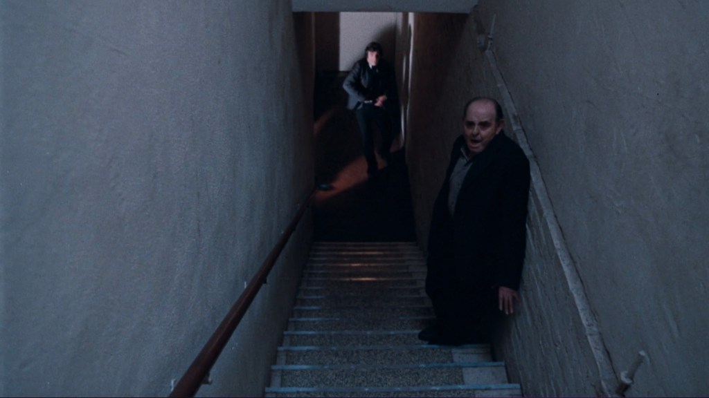 A dimly lit stairwell featuring a man standing at the top and another man walking down the stairs, creating an atmosphere of tension.