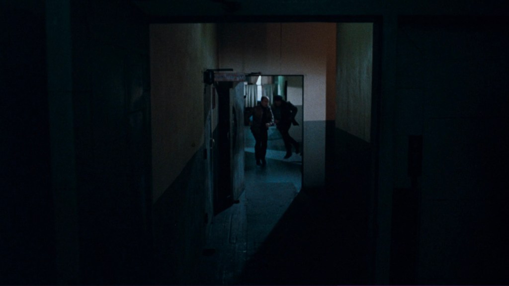 A shadowy corridor with two figures hurriedly exiting a door in the background, surrounded by dim lighting.