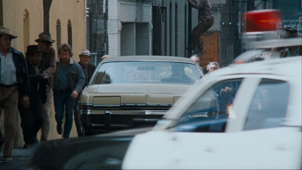 A scene depicting a chaotic urban street with a crowd of people running towards a car, while police vehicles are chasing them in the foreground.