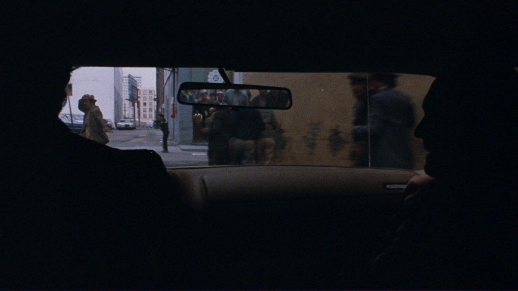 View from inside a car showing two silhouettes of people in the front seat, with a city street visible through the windshield, featuring pedestrians and buildings in the background.
