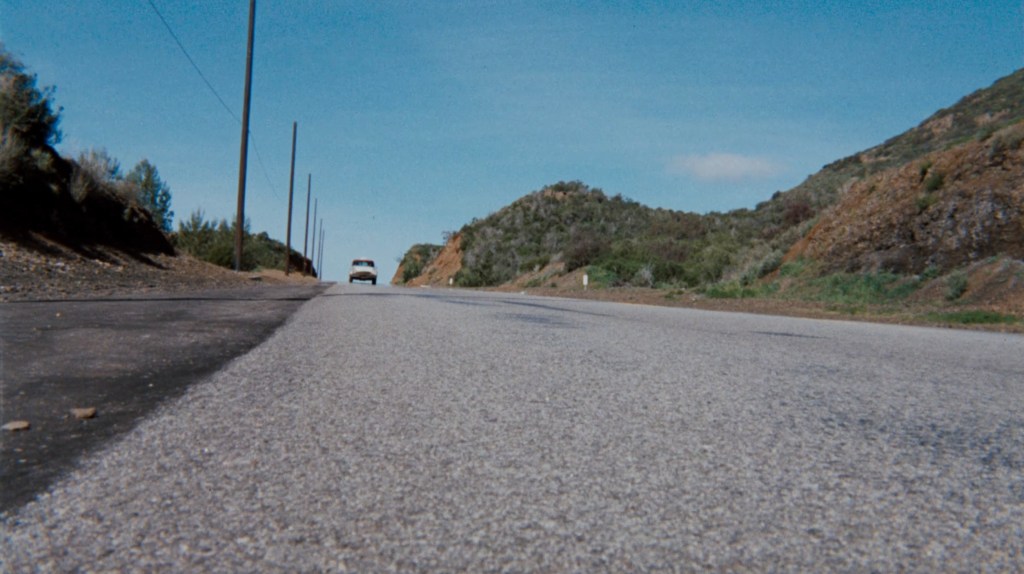 A view of a road stretching into the distance with a vehicle approaching, flanked by green hills and blue sky.