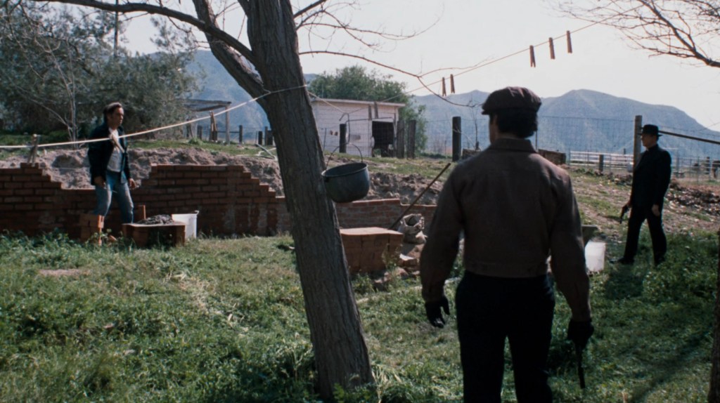A tense standoff in a rustic outdoor setting, featuring a woman on the left and two men in front of her, one holding a weapon. The background includes a tree, a brick wall, and distant mountains.
